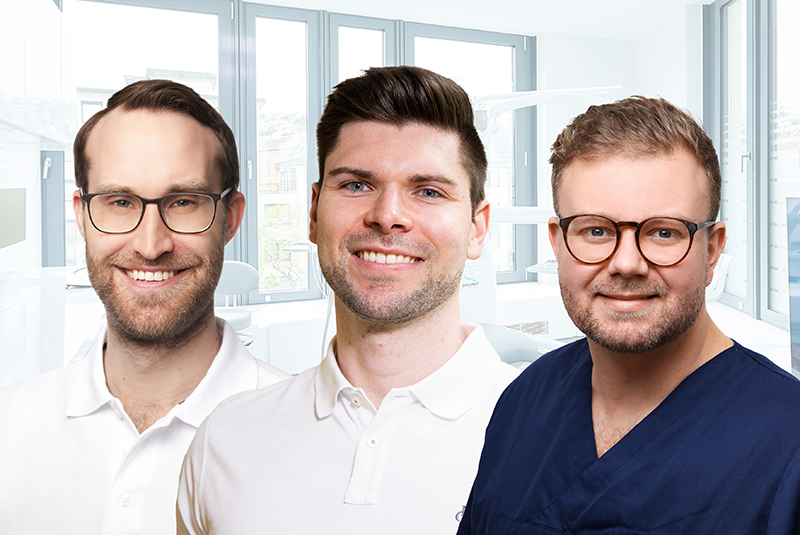 Drei Zahn&auml;rzte stehen gemeinsam in einer modernen Zahnarztpraxis und l&auml;cheln in die Kamera